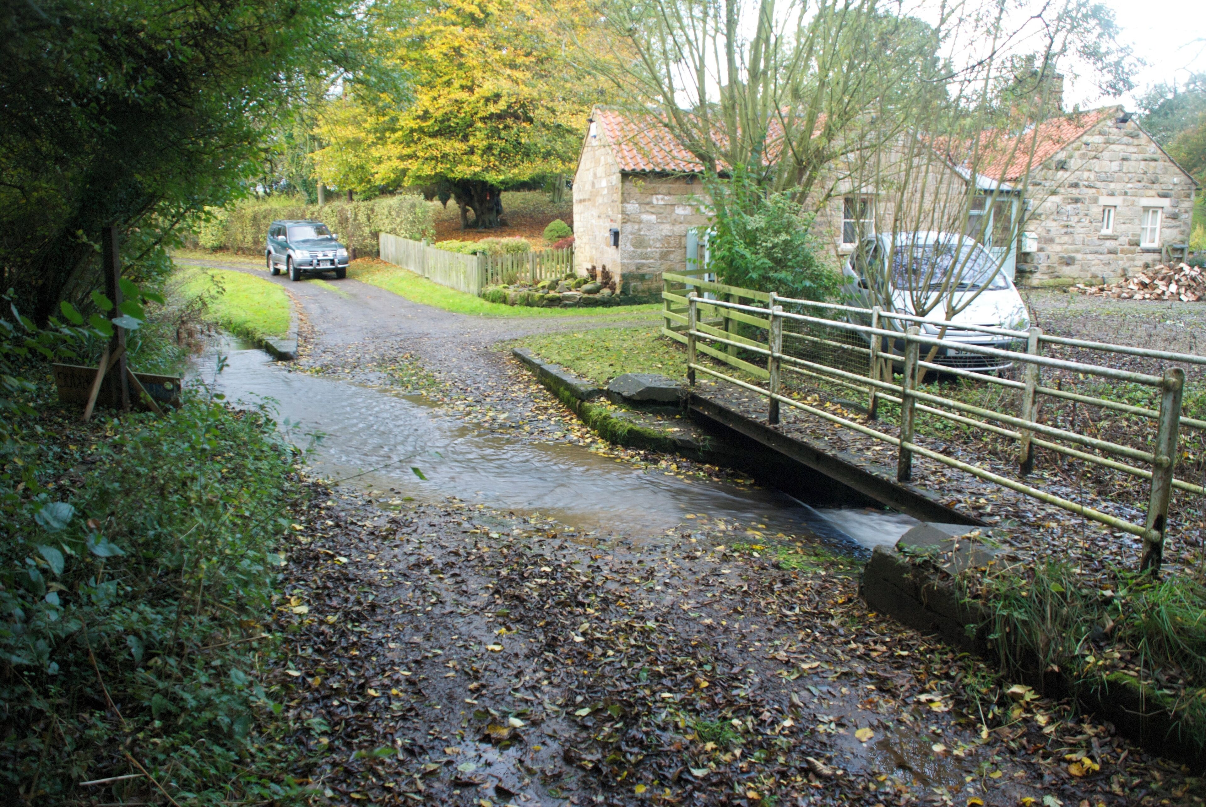 A rural road with a stone bridge, a stone house, and parked cars.