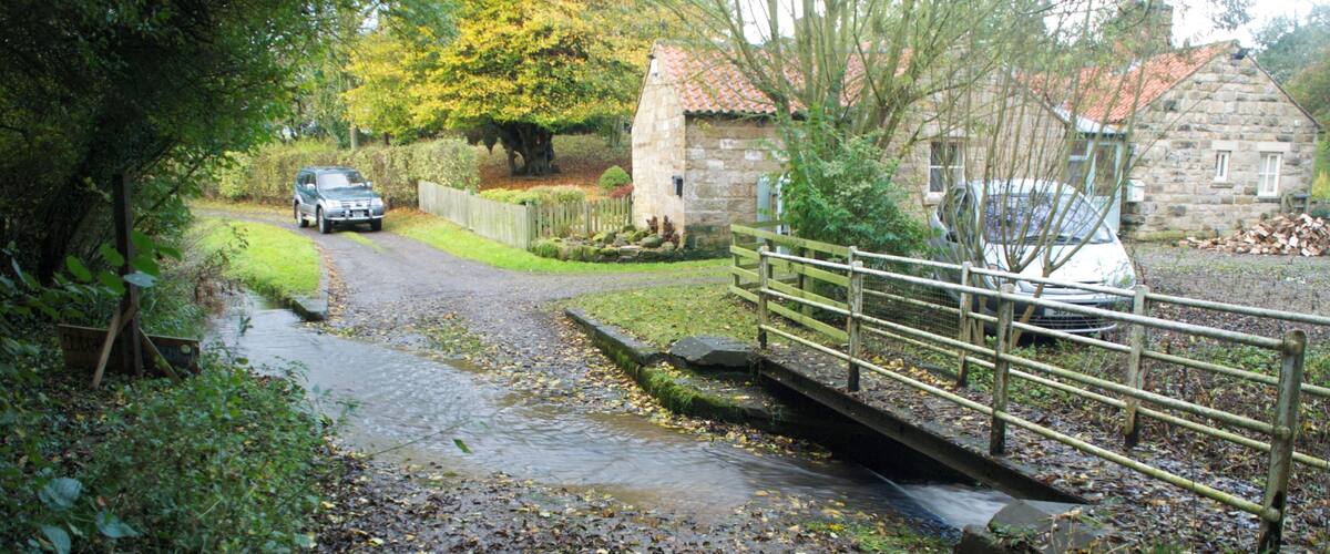 A rural road with a stone bridge, a stone house, and parked cars.