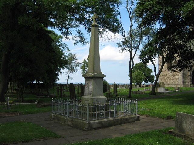 A green obelisk monument surrounded by a metal fence in a cemetery with trees and a stone building in the background.