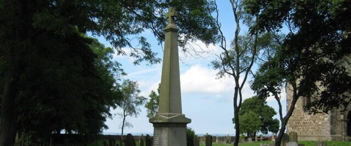 A green obelisk monument surrounded by a metal fence in a cemetery with trees and a stone building in the background.