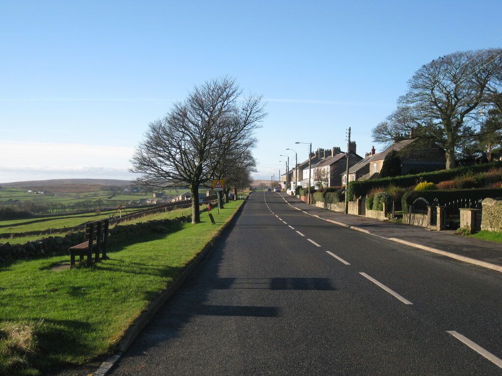 A rural road flanked by houses and trees, with a clear sky and distant hills.