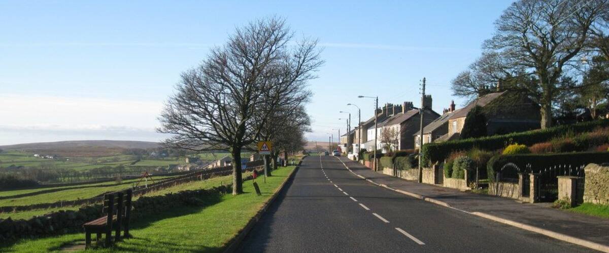 A rural road flanked by houses and trees, with a clear sky and distant hills.