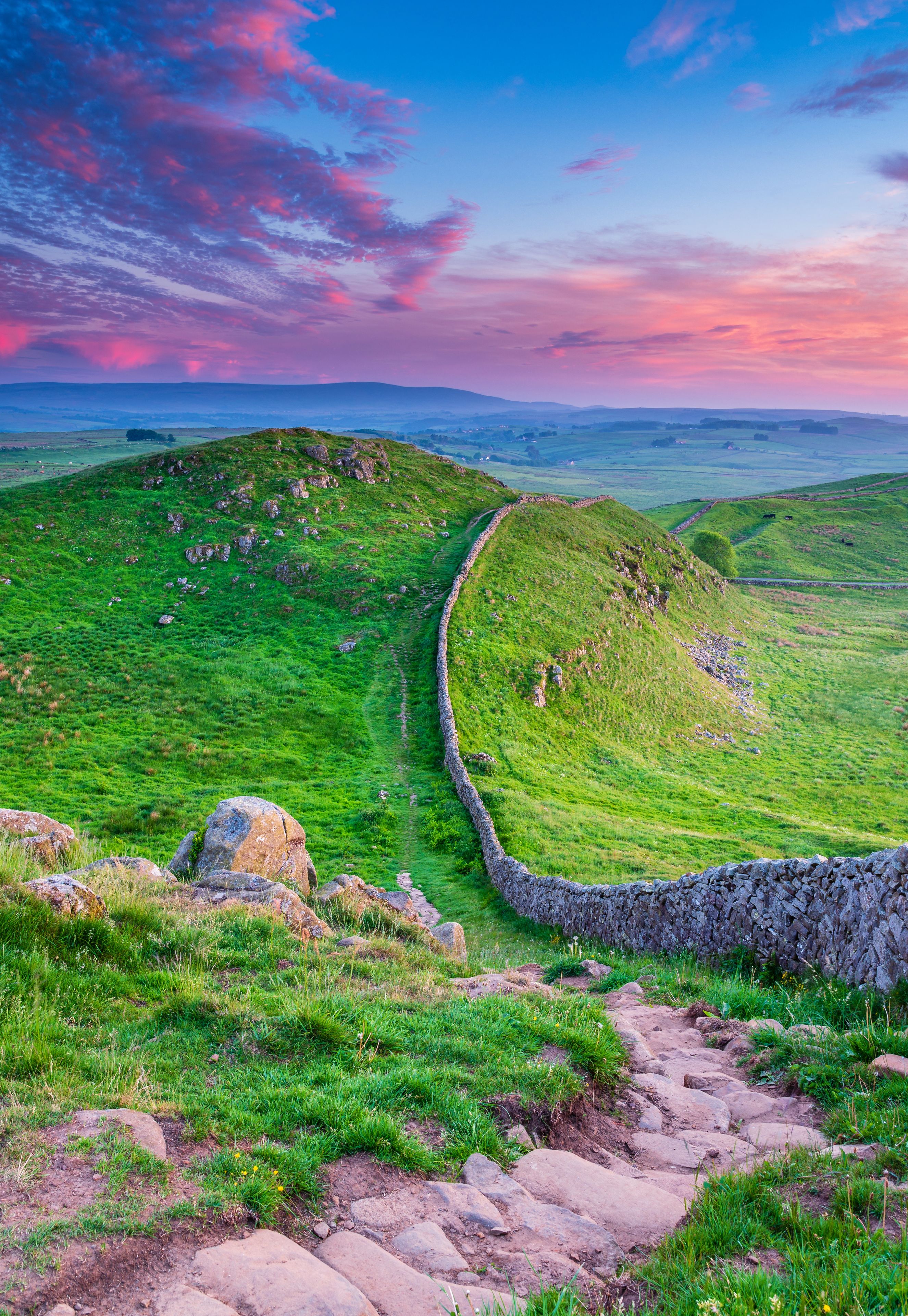A lush green landscape with a stone wall meandering through rolling hills, under a vibrant sunset sky.