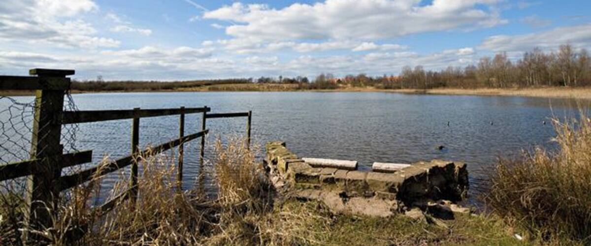 A calm lake with a wooden fence and a stone structure in the foreground.