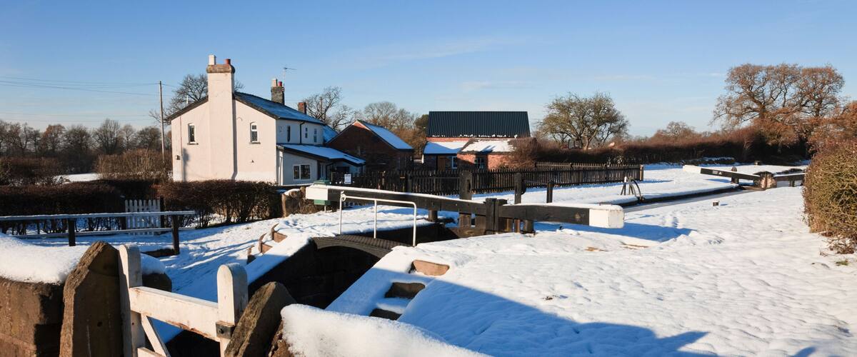 A snow-covered landscape with a stone bridge, wooden fence, and houses in the background.