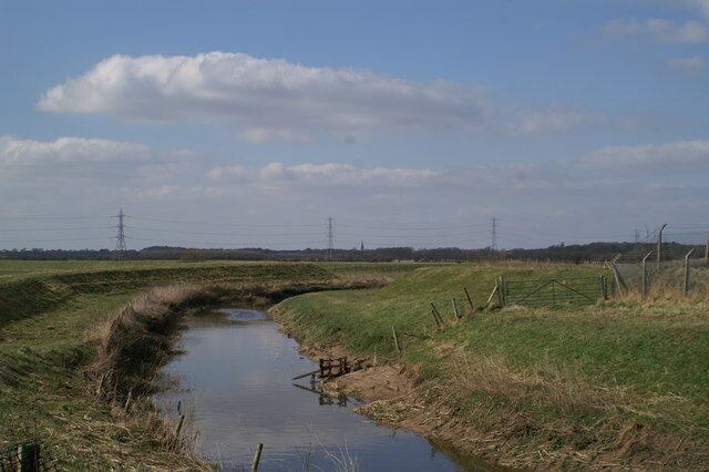 A narrow water channel in a rural landscape with grassy banks and distant power lines.
