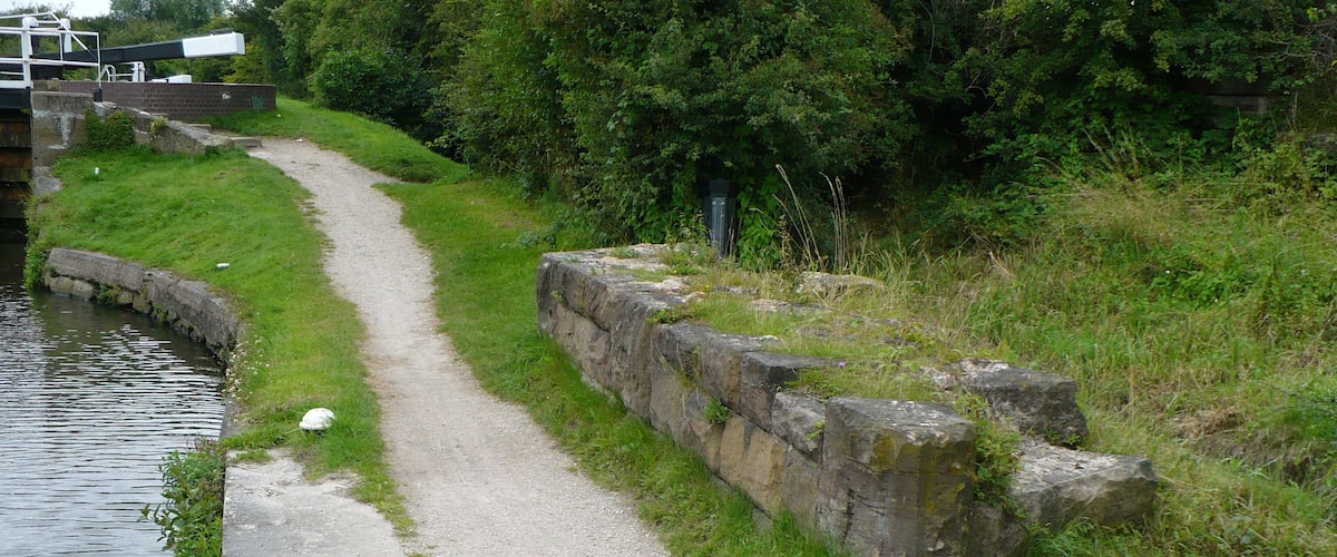 A narrow concrete path beside a canal, bordered by grass and stone walls.