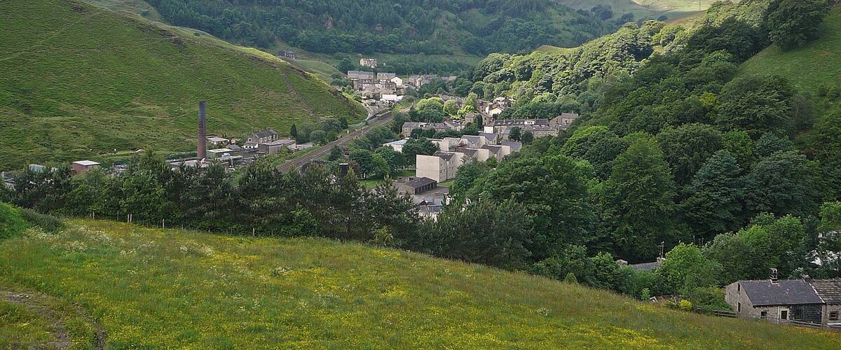 A valley with a small town nestled among green hills, a railway line, and a tall chimney.