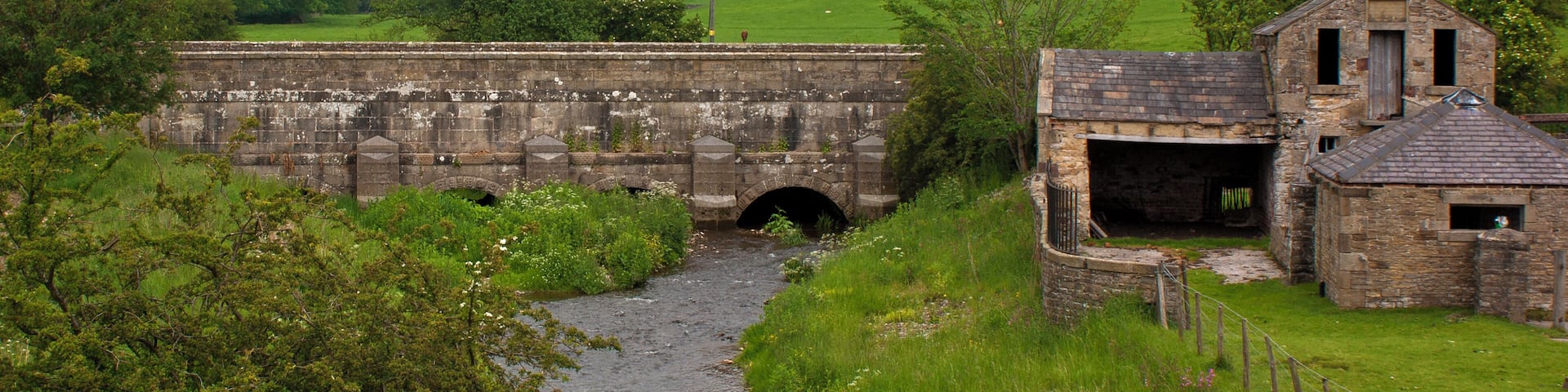 A stone bridge over a river, surrounded by lush greenery and a small stone building.
