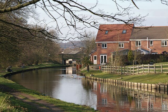 A calm river with a wooden fence, a brick house, and a bridge in the background.