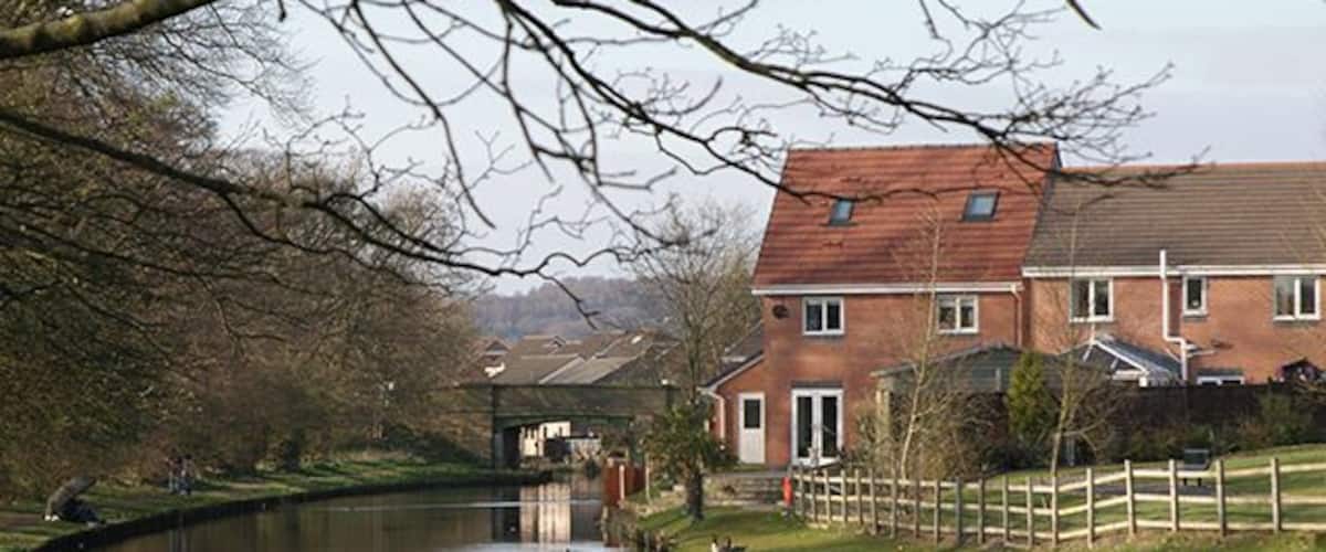 A calm river with a wooden fence, a brick house, and a bridge in the background.