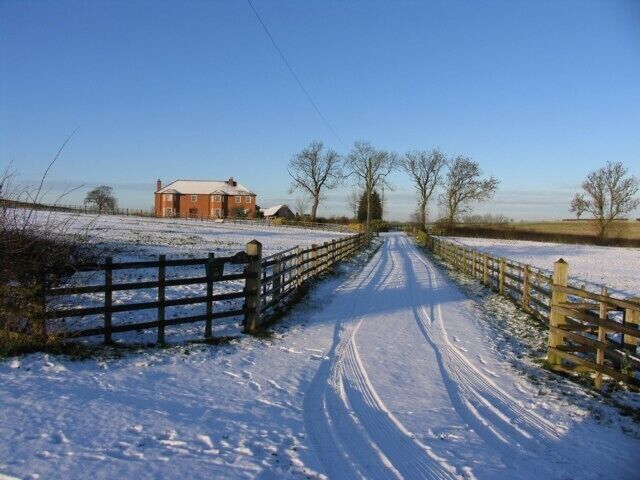 A snow-covered rural road with a wooden gate, a brick house, and open fields.