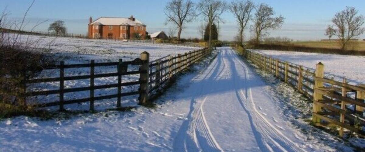 A snow-covered rural road with a wooden gate, a brick house, and open fields.