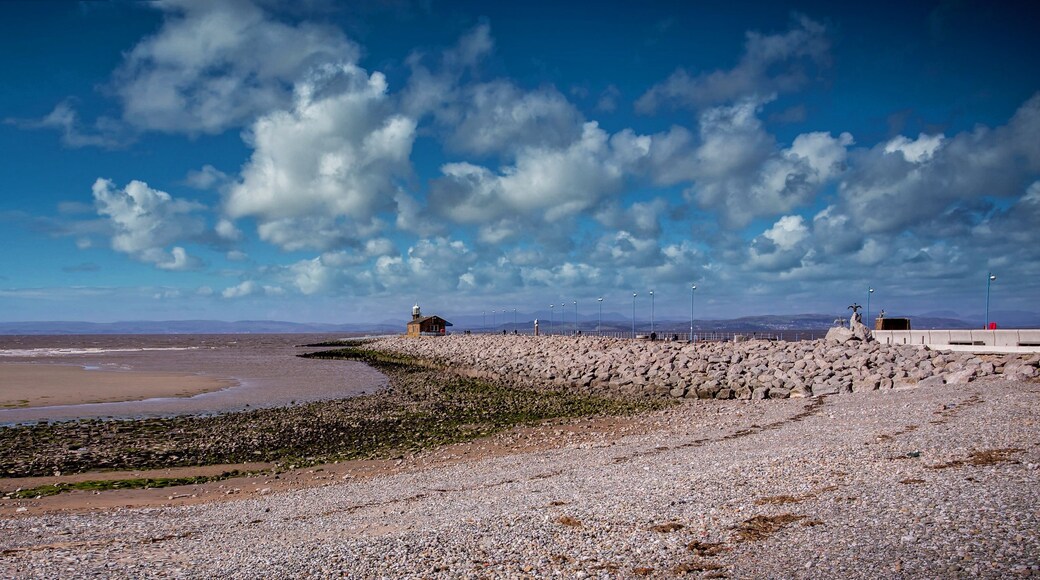 A coastal landscape with a rocky shoreline, a lighthouse, and distant mountains under a partly cloudy sky.