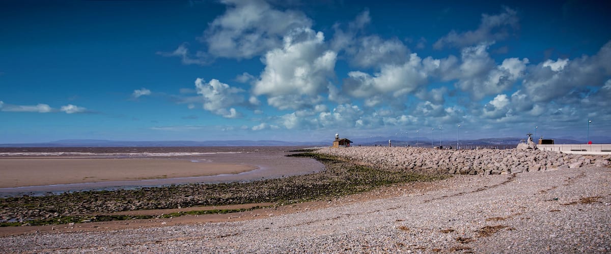 A coastal landscape with a rocky shoreline, a lighthouse, and distant mountains under a partly cloudy sky.