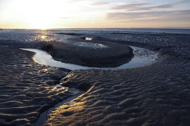 A wet sand beach at sunset with a small water channel and ripples.