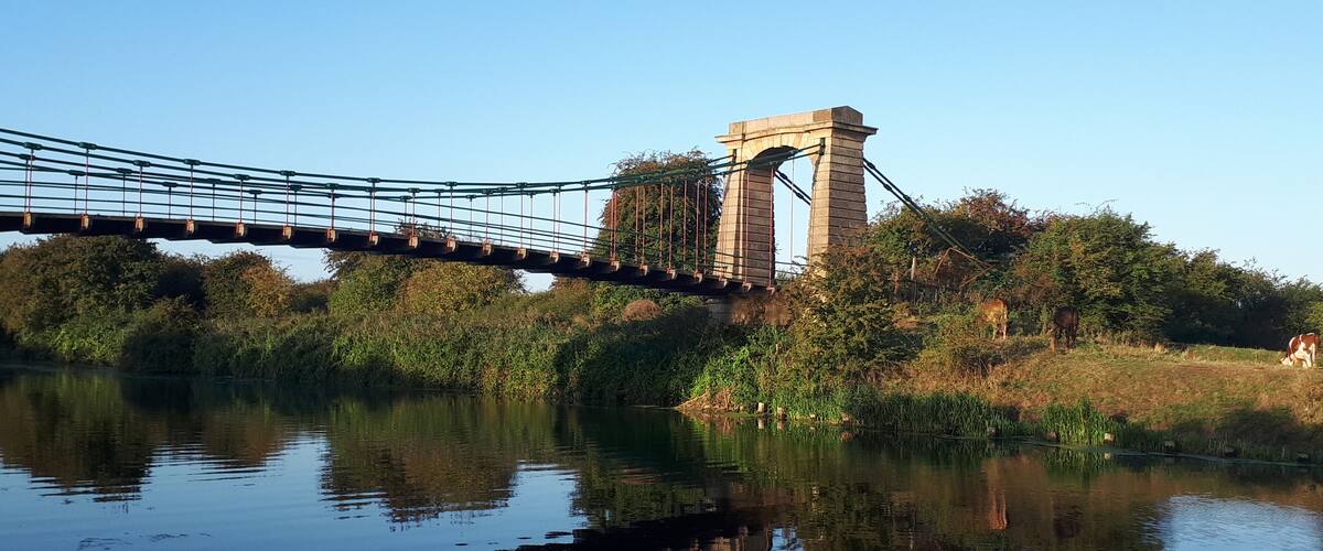 A suspension bridge over a calm river with lush greenery and trees on either side.