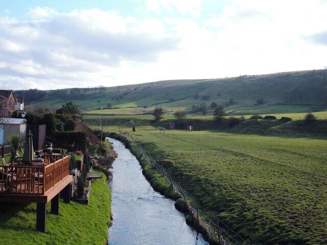 A river flowing through a green valley with a wooden deck and buildings on the left.