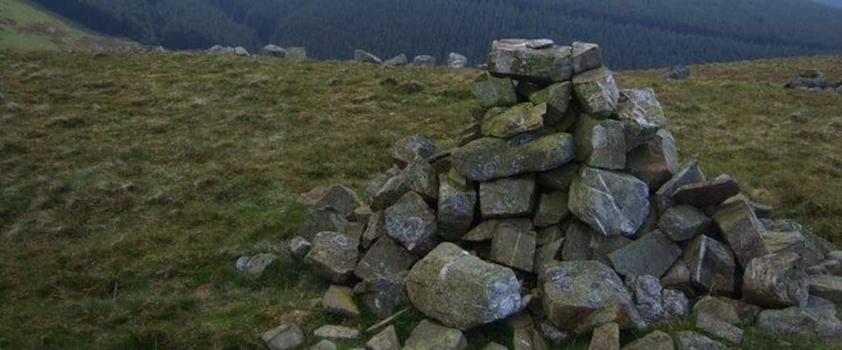 A stone cairn on a grassy hill with a forested valley in the background.