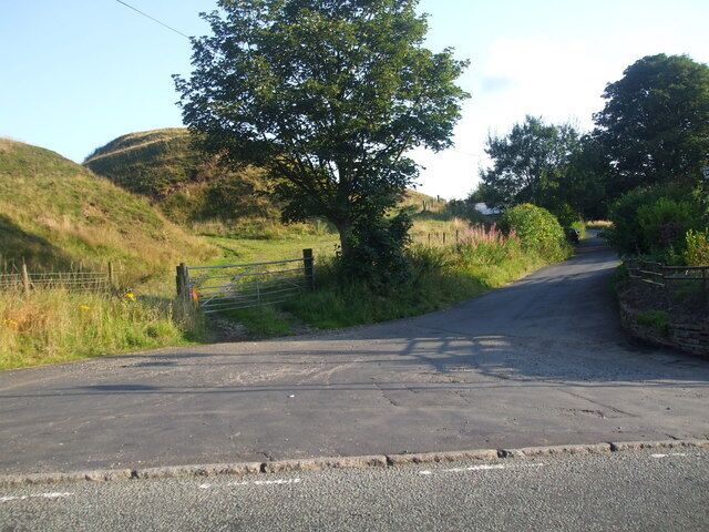 A rural road with a stone wall and a metal gate, surrounded by greenery and hills.