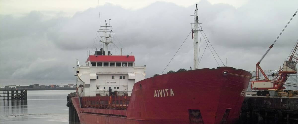 A large red ship named 'AVITA' docked at a port with cranes and a white building in the background.