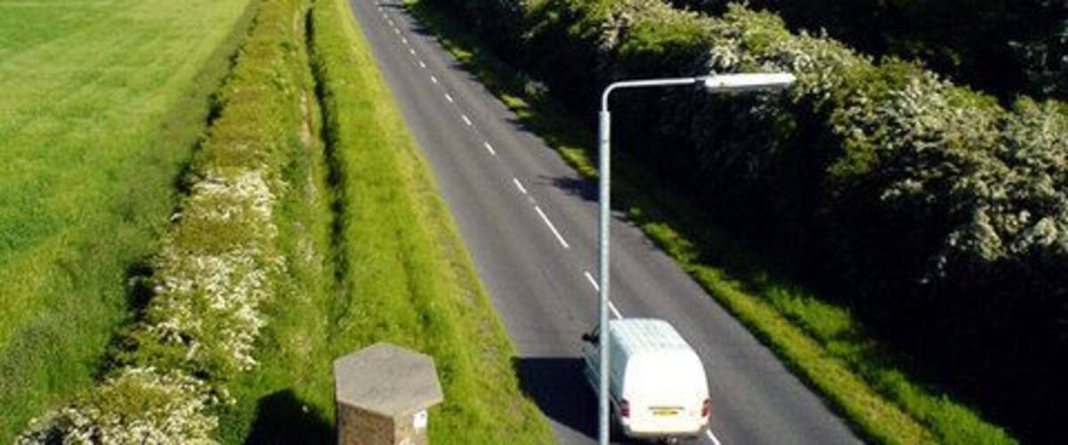 A rural road with a white van, surrounded by green fields and hedgerows.