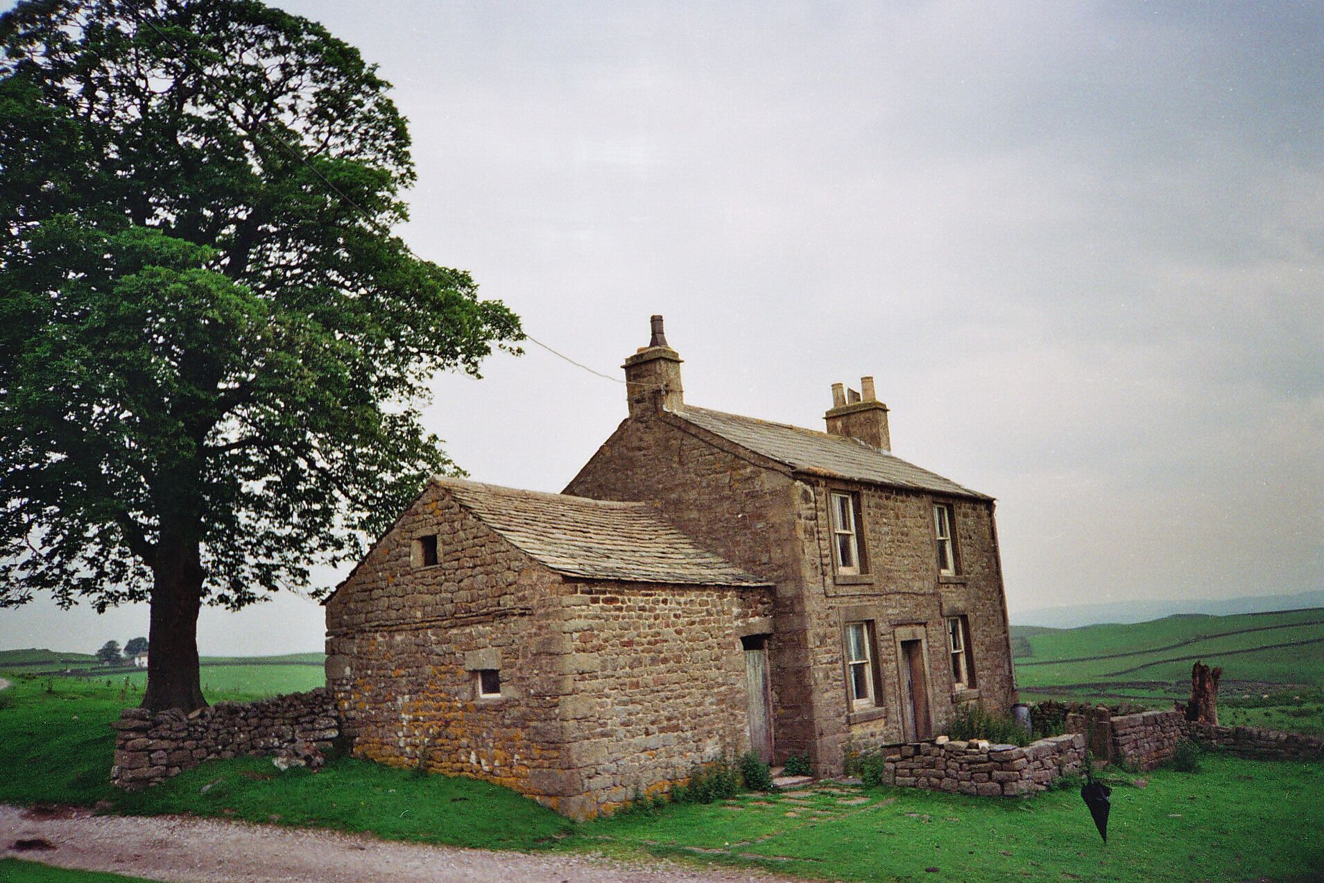 A stone house with a sloped roof, surrounded by a stone wall and a large tree.