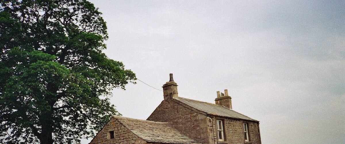 A stone house with a sloped roof, surrounded by a stone wall and a large tree.