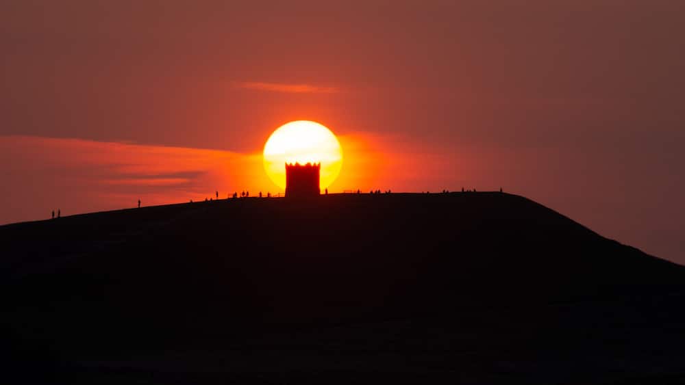 Ein Sonnenuntergang über einer Hügelkulisse mit der Silhouette eines Turms und Personen auf der Spitze.