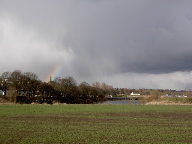 A rural landscape with a rainbow, a body of water, and houses in the background.