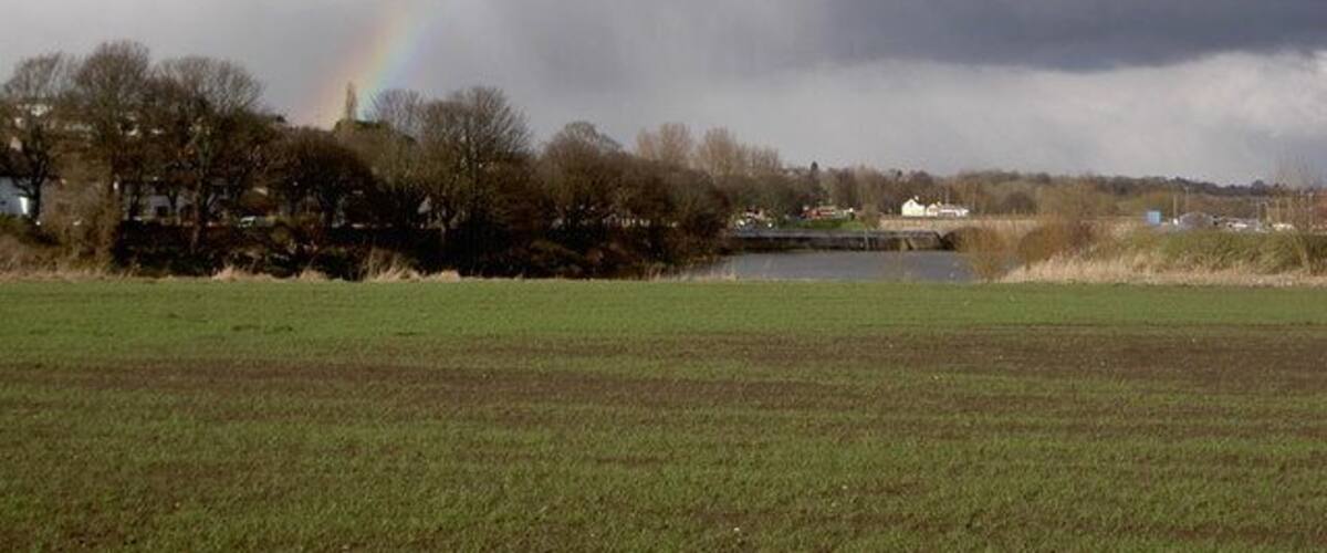 A rural landscape with a rainbow, a body of water, and houses in the background.