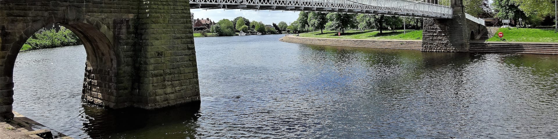 A stone bridge with a metal arch over a river, surrounded by greenery and a clear sky.