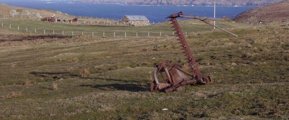 A coastal landscape with a rusty metal structure, a fence, and a distant view of the sea and hills.