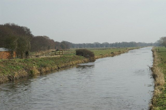 A narrow waterway flanked by grassy banks and trees, with a wooden fence and a small building in the distance.