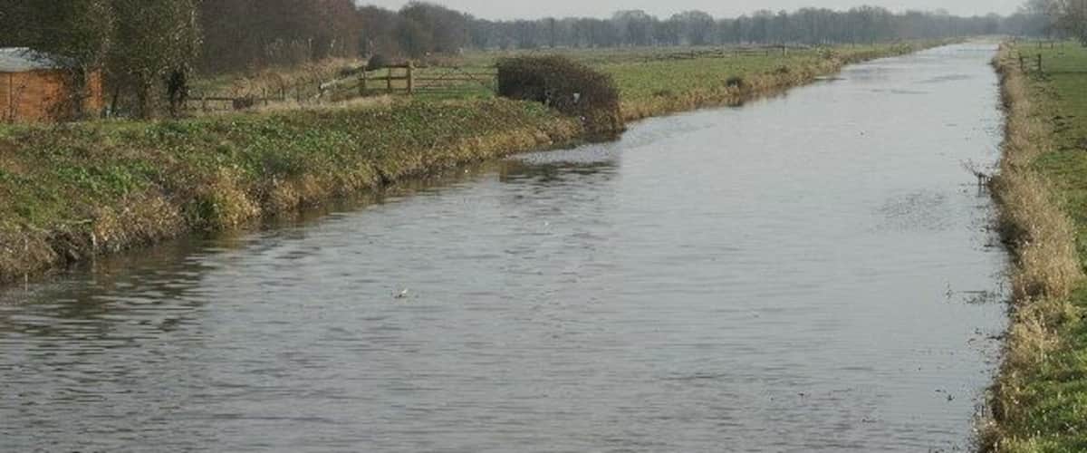 A narrow waterway flanked by grassy banks and trees, with a wooden fence and a small building in the distance.