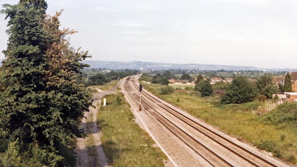 A railway track with multiple lines, surrounded by greenery and residential buildings in the distance.