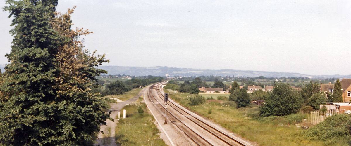 A railway track with multiple lines, surrounded by greenery and residential buildings in the distance.