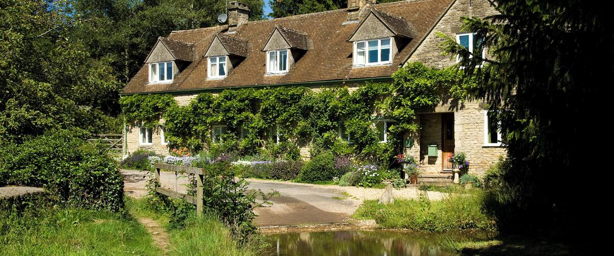 A stone house with multiple gabled roofs, a pond in the foreground, and lush greenery surrounding the property.