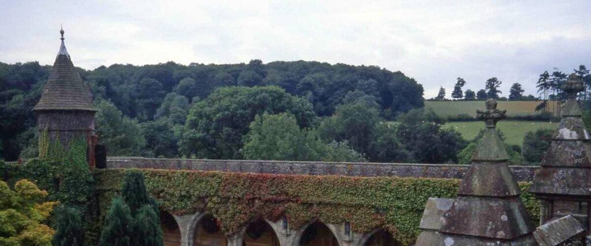 A historic cloister with arches, a tower, and a lush green landscape in the background.