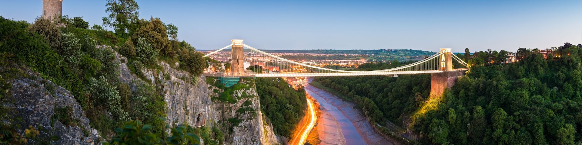 A suspension bridge over a river, surrounded by dense forest and a historic tower.