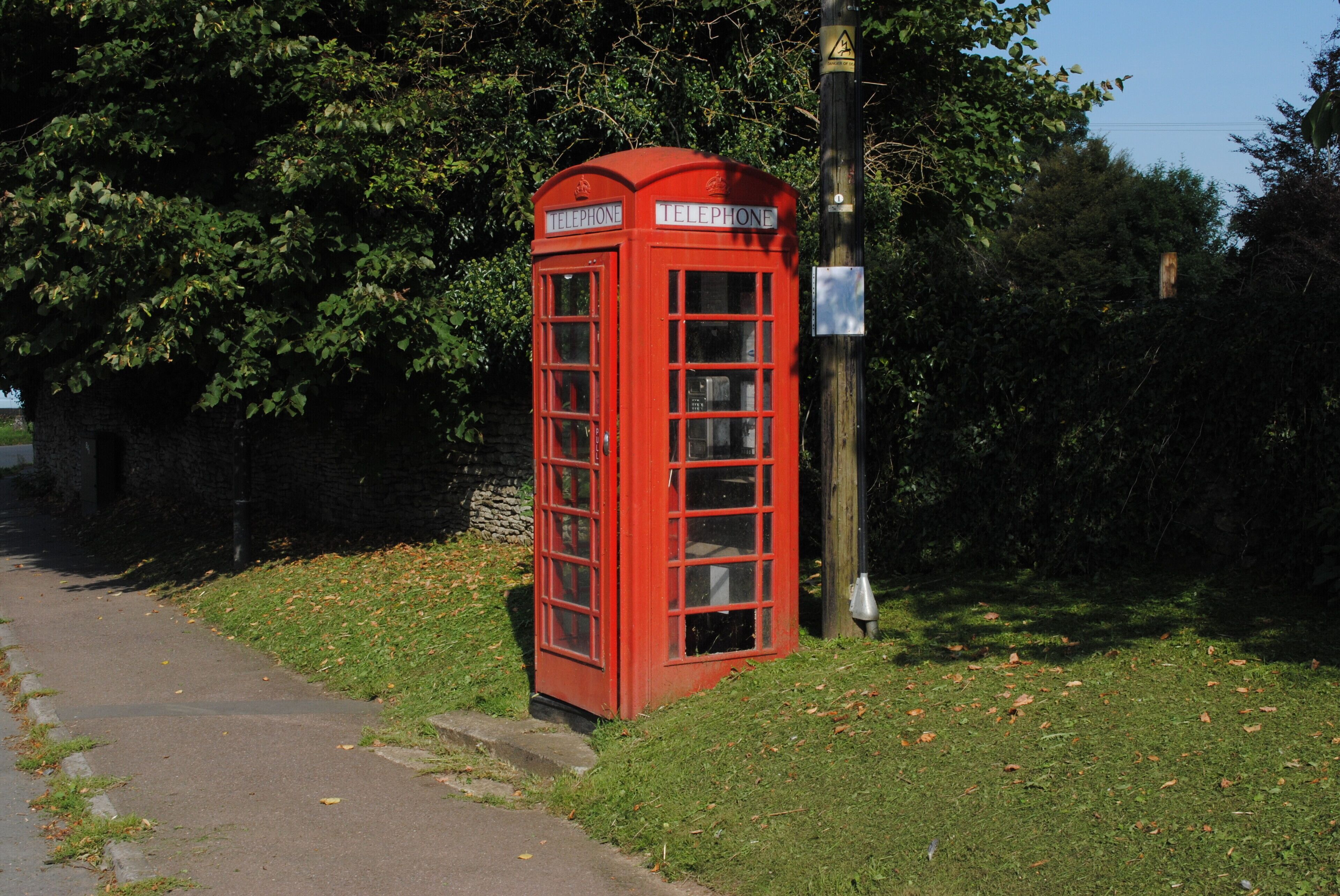 A red telephone booth on a grassy path with trees in the background.