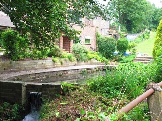 Moddershall Mill. A modern house stands downstream from the original mill which can be seen in the right background. The mill stream has been converted into an attractive water feature.