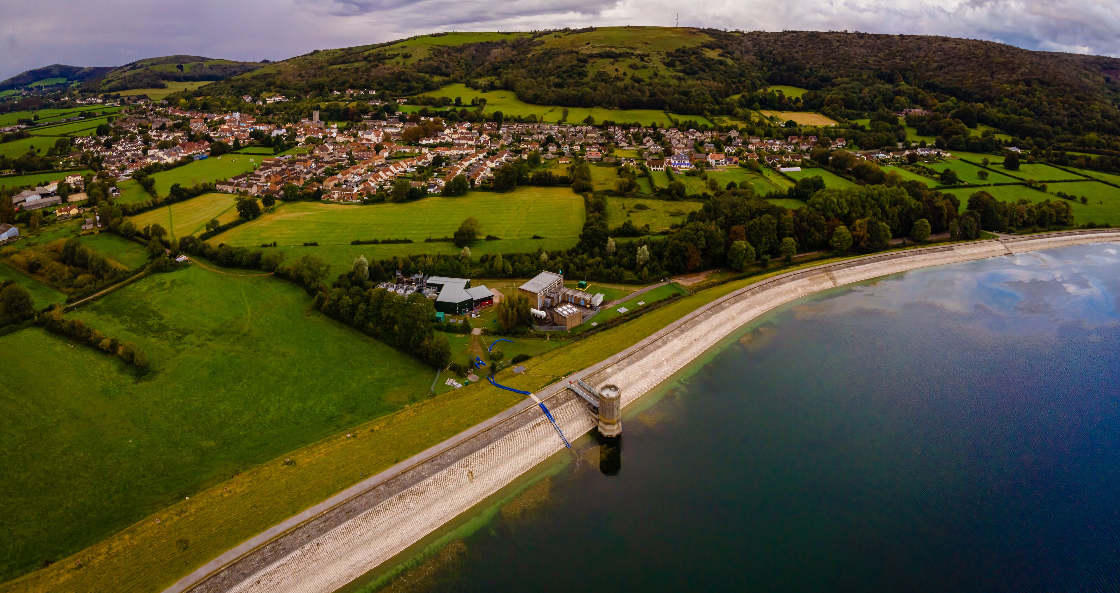 An aerial view of a coastal town with a bridge connecting to a peninsula, surrounded by green fields and hills.