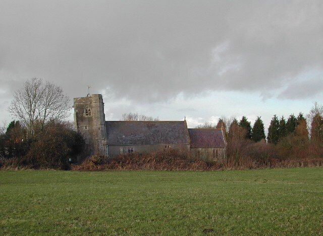 A church with a bell tower and a stone building surrounded by a grassy field and trees.