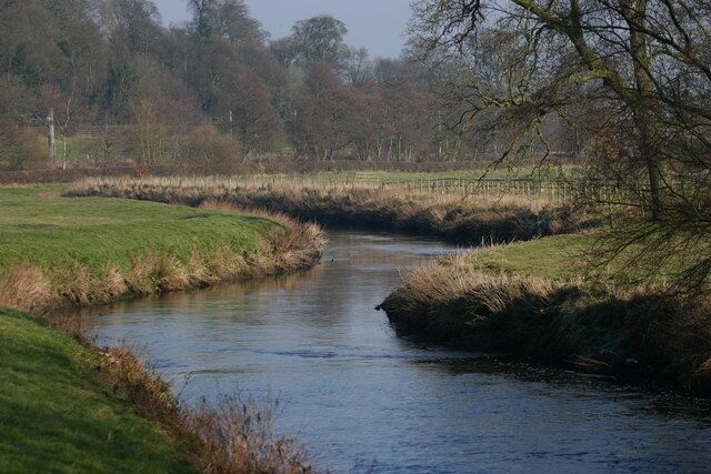 A calm river meandering through a grassy landscape with trees and a fence in the background.