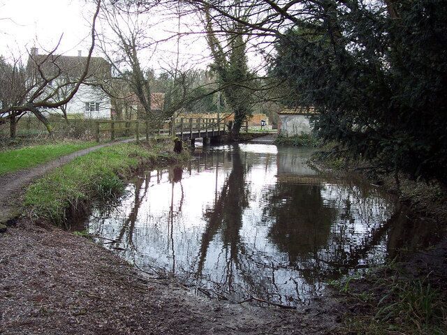 A calm river with a wooden bridge, surrounded by trees and a grassy path.