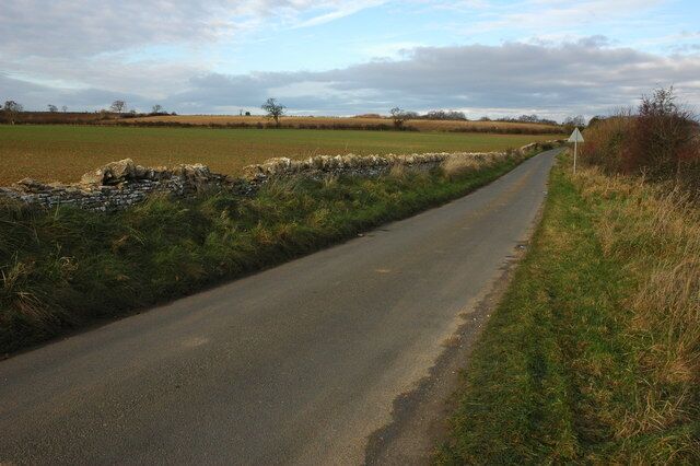 A narrow rural road flanked by stone walls and fields under a partly cloudy sky.