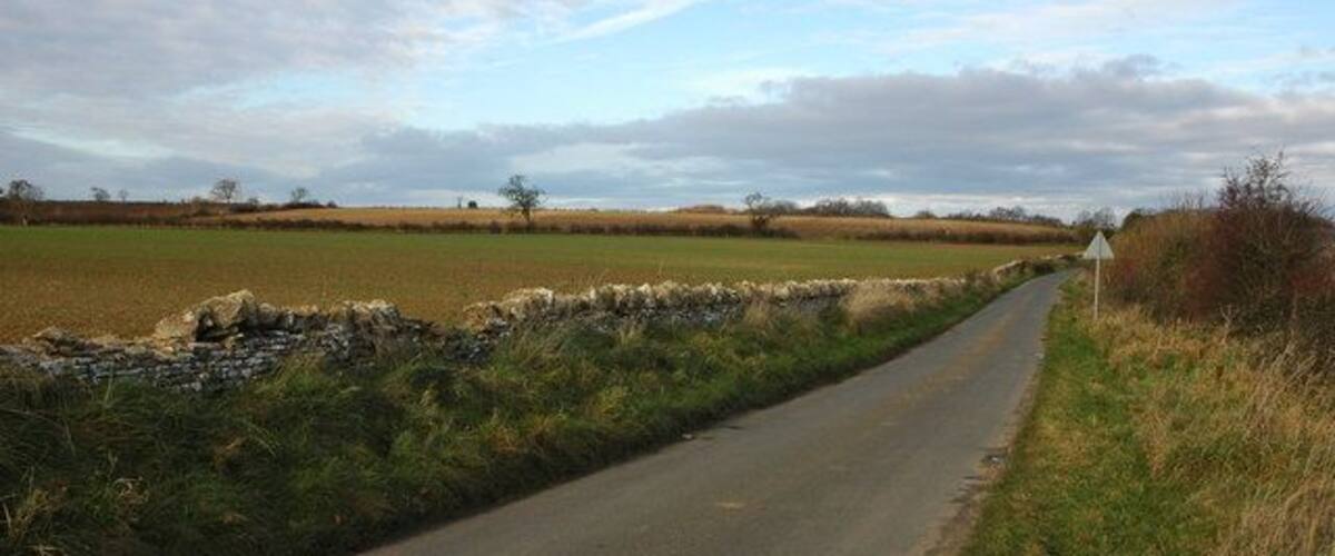 A narrow rural road flanked by stone walls and fields under a partly cloudy sky.