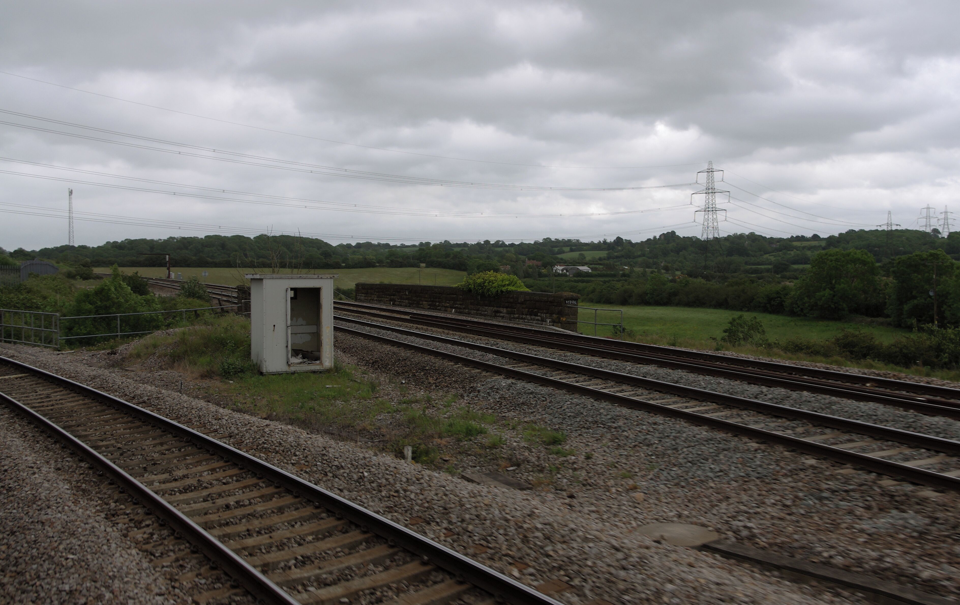 Railway tracks, a small utility box, and a rural landscape with trees and power lines.