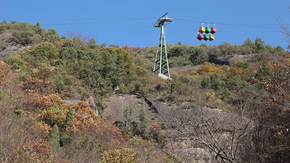 A cable car system with colorful gondolas against a backdrop of autumn-colored trees and rocky terrain.